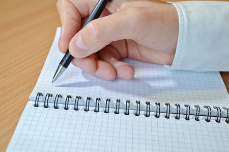 A man's hand in a white shirt writes the text with a black pen in a notebook with a spiral on a wooden table closeupの写真素材