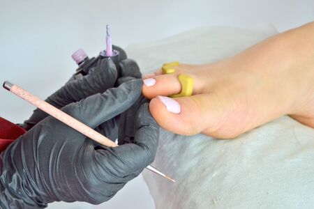 A pedicure master in black gloves paints the nails of the girl feet with light pink nail polish with a brushの写真素材