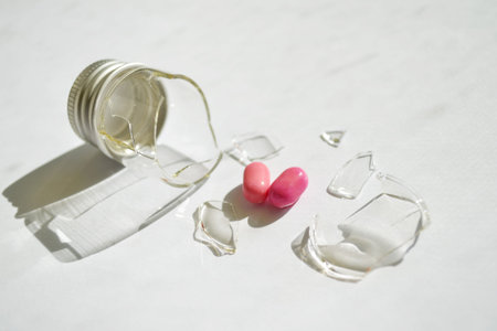 A broken small glass bottle with a metal cap, two pink heart-shaped tablets fell out of it, on a...