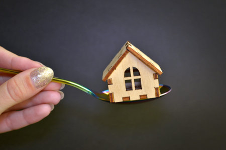 Wooden yellow toy house lies on a colored metal spoon held by a female hand with manicure on a black...