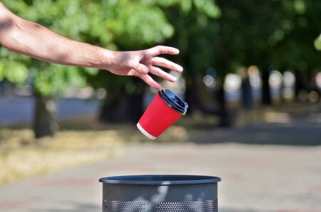 Male hand threw a red paper cup for coffee in a trash can, a cup flies in the air, against a background of green treesの写真素材