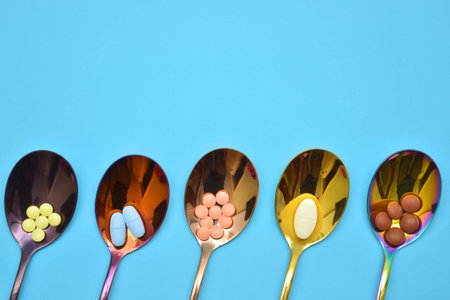 Five different types of pills in five multi colored metal spoons on a blue background with copyspace