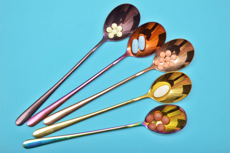 Five different types of pills in five multi colored metal spoons with long handles on a blue...