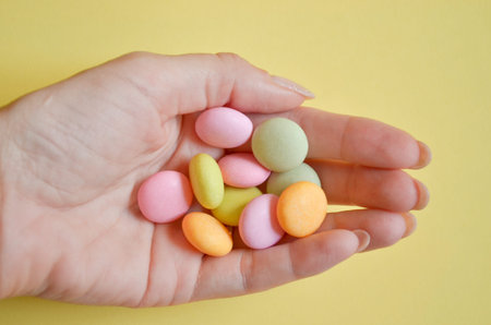 Round multicolored candies lie in bunch on woman hand on yellow color, closeup