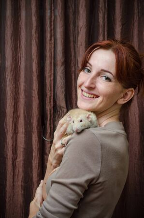 Happy smiling redhaired girl holding a light rat in her arms on a brown background, a symbol of the new year, vertically orientedの写真素材