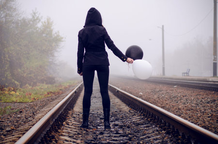 Lonely girl in black clothing standing between the rails in a fog. She holds a black and white...