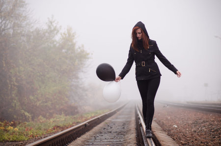 Lonely girl in black clothing walking on the rails in a fog. She holds a black and white balloon in...