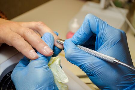 Manicure procedure. The master in blue gloves cleans the skin around the nail and moves the cuticle with a pusherの写真素材