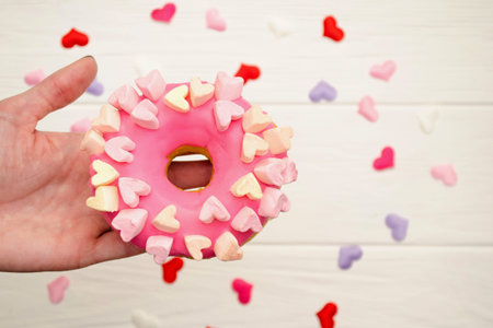 Sweet pink donut with icing and decoration with hearts on a rainbow wooden background in female hand