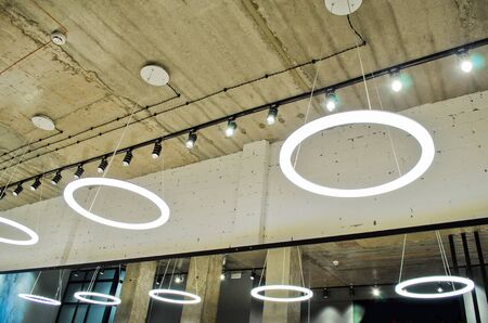 White circular ring shaped lamps hang under a concrete ceiling with external wiring and black fixtures, with mirror reflectionの写真素材