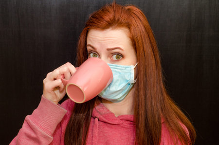 Young caucasian redhaired girl with green eyes, in a medical mask in a pink sweater on a black...