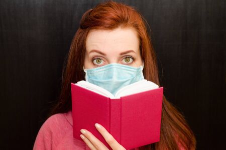 Young caucasian redhaired girl with green eyes, read book in a medical mask in a pink sweater on a black background, coronavirus epidemic, quarantineの写真素材