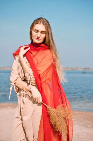 Young beautiful caucasian blond woman in a light boho dress and a red thin scarf, stays on beach by water under sunshineの写真素材