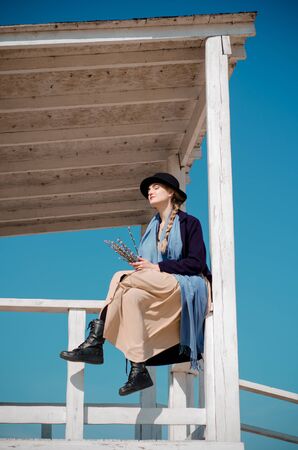 Young caucasian blonde girl in a boho dress, coat and hat in spring on city beach, in lifeguard house under rays of spring sun with willow branches, smilingの写真素材