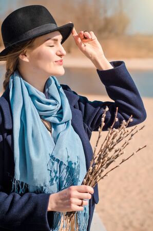 Young caucasian blonde girl in a boho dress, coat and hat in spring on beach, under rays of spring sun with willow branches, smilingの写真素材