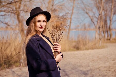 Young caucasian blonde girl in a boho dress, coat and hat in spring on beach, under rays of spring sun with willow branches, smiling dream about somethingの写真素材