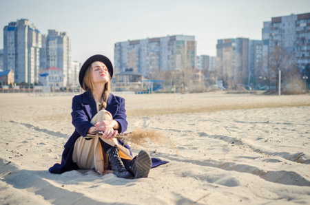 Young caucasian blonde girl in light dress, coat and hat on beach, under rays of spring sun with...