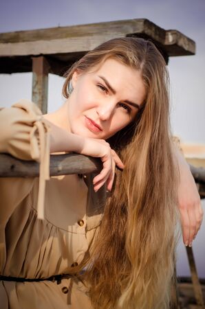 Beautiful young caucasian blonde girl, in a light boho dress, sits on an old wooden construction, a bridge over the water against blue sky in countrysideの写真素材