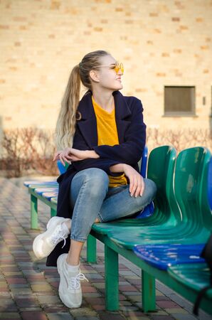 Beautiful young blonde girl in a yellow sweater and blue jeans, white sneakers and a blue coat sits on the seats for spectators on the sports field in yellow glassesの写真素材