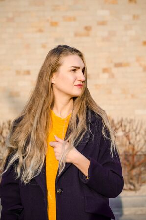 Beautiful young blonde girl in a yellow sweater and blue jeans, white sneakers and a blue coat, in yellow glasses stay against a brick wallの写真素材