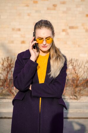 Beautiful young blonde girl in a yellow sweater and blue jeans, white sneakers and a blue coat, in yellow glasses speaks on the phone against a brick wallの写真素材