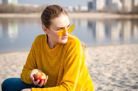 Beautiful young blonde girl in a yellow sweater and blue jeans, stay on beach against a blue sky and water in yellow glasses and hold red appleの写真素材