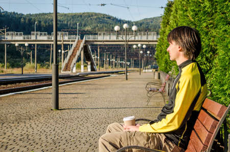 Young man in sport tourist clothes sits on the platform of the station, near the rails, and holds cup of coffeeの写真素材