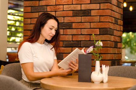 Young happy european girl with brown hair sitting in a cafe near a brick wall reading a book at the...
