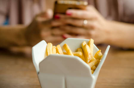 Fries are served at the restaurant in a in white porcelain box styled like take away box. On a wooden table on the background of female hands with a phone on lunchの写真素材