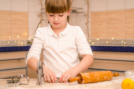 Cute happy girl learns how to cook Christmas gingerbread cookies in a bright cozy kitchen, kneads the dough and rolls it out with a rolling pin, cuts cookies with moldsの写真素材