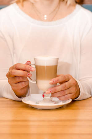 Young woman in a cafe drinks a latte, holds a transparent glass with her hand on a white saucer. Waits for the coffee to cool. With copyspaceの写真素材