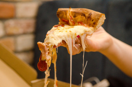 Very cheesy pizza slice with pepperoni in female hand, melted cheese stretches in long strands, in a cafe against background of a box, sofa and a brick wallの写真素材