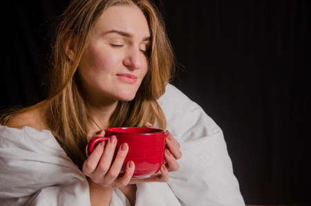 Beautiful young woman with blonde hair, sits in the morning wrapped in a white blanket on black background, drinks tea from a red cupの写真素材