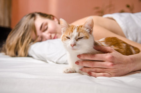 Beautiful young woman lies in a white bed with a ginger cat, smiles, kisses and hugs her, love animals, friendshipの写真素材