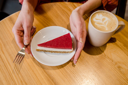 Young woman at table in cafe eats red cake on saucer with her hands and a fork, there is cup with...
