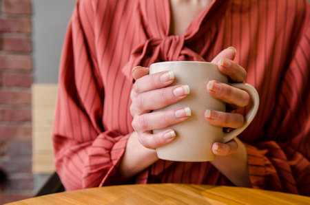 Young female hands holding and hugging a cup of coffee with latte art at a wooden table in a cafe. woman in red dressの写真素材