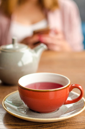 Red cup of red tea, a gray teapot stand on a table in a cafe. In the background, a young woman is...