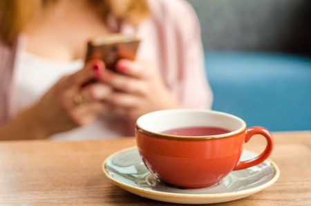 Red cup of red tea, a gray teapot stand on a table in a cafe. In the background, a young woman is working on her smartphoneの写真素材