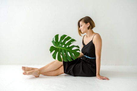 Young European woman in a black long dress, sitting on the floor, holding a large monstera leaf in her hands on a white light room backgroundの写真素材