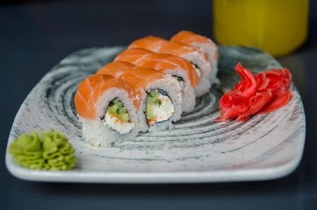 Appetizing portion of sushi rolls philadelphia with red fish salmon, with soft cheese, cucumbers, avocado and ginger with wasabi, on a gray plate on a black table, on background of yellow fruit juiceの写真素材
