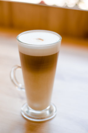 Beautiful tall transparent glass with a handle containing a latte coffee drink on a table, in a cafe in front of a window with natural lightの写真素材