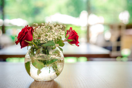 Street summer cafe terrace, with white and wooden furniture, transparent glass round vase with red roses on table, blurred background without peopleの写真素材