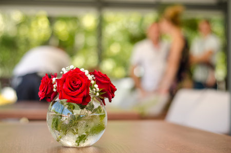 Street summer cafe terrace, with white and wooden furniture, transparent glass round vase with red roses on table, blurred background with peopleの写真素材