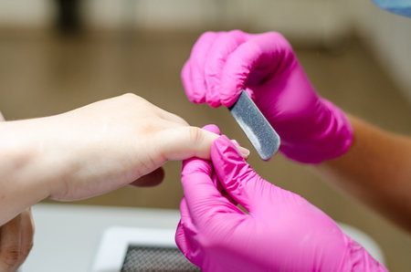 Female manicurist in pink gloves works with young hands and nails under a bright lamp. Process of sawing nails with a sawの写真素材