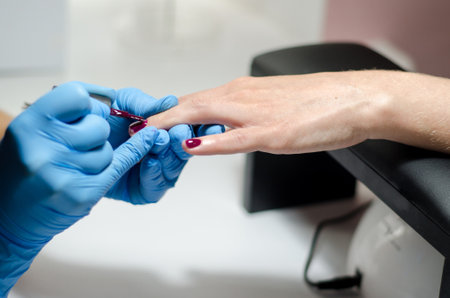 Female manicurist in blue gloves works with young hands and nails under a bright lamp. Process of applying gel polish on nailsの写真素材