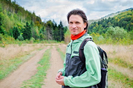 Handsome young smiling male tourist in green jacket and orange scarf, with dark hair and bzckpack, standing on a path through the mountains, enjoying the viewの写真素材