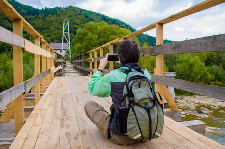 Young man tourist in a green jacket and with a backpack sits on a wooden bridge near the mountains, and takes pictures of them on a smartphoneの写真素材