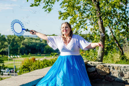 Street performer, beautiful young woman in blue skirt and white blouse, with soap bubbles shows soap show in summer in open air park for childrenの写真素材