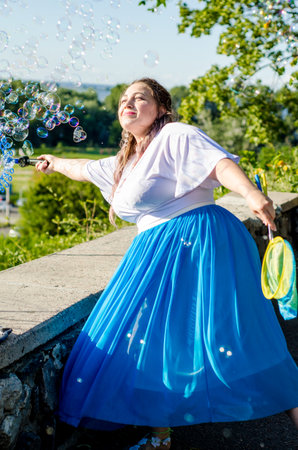 Street performer, beautiful young woman in blue skirt and white blouse, with soap bubbles shows soap show in summer in open air park for childrenの写真素材