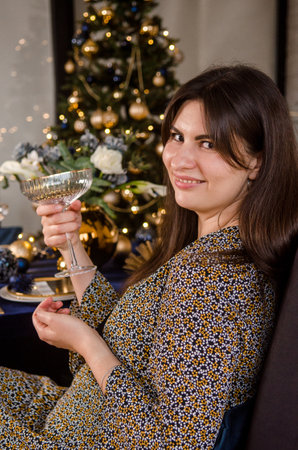 Young European woman with dark hair is sitting at a festive New Year table with a glass of sparkling wine, against the background of a Christmas treeの写真素材